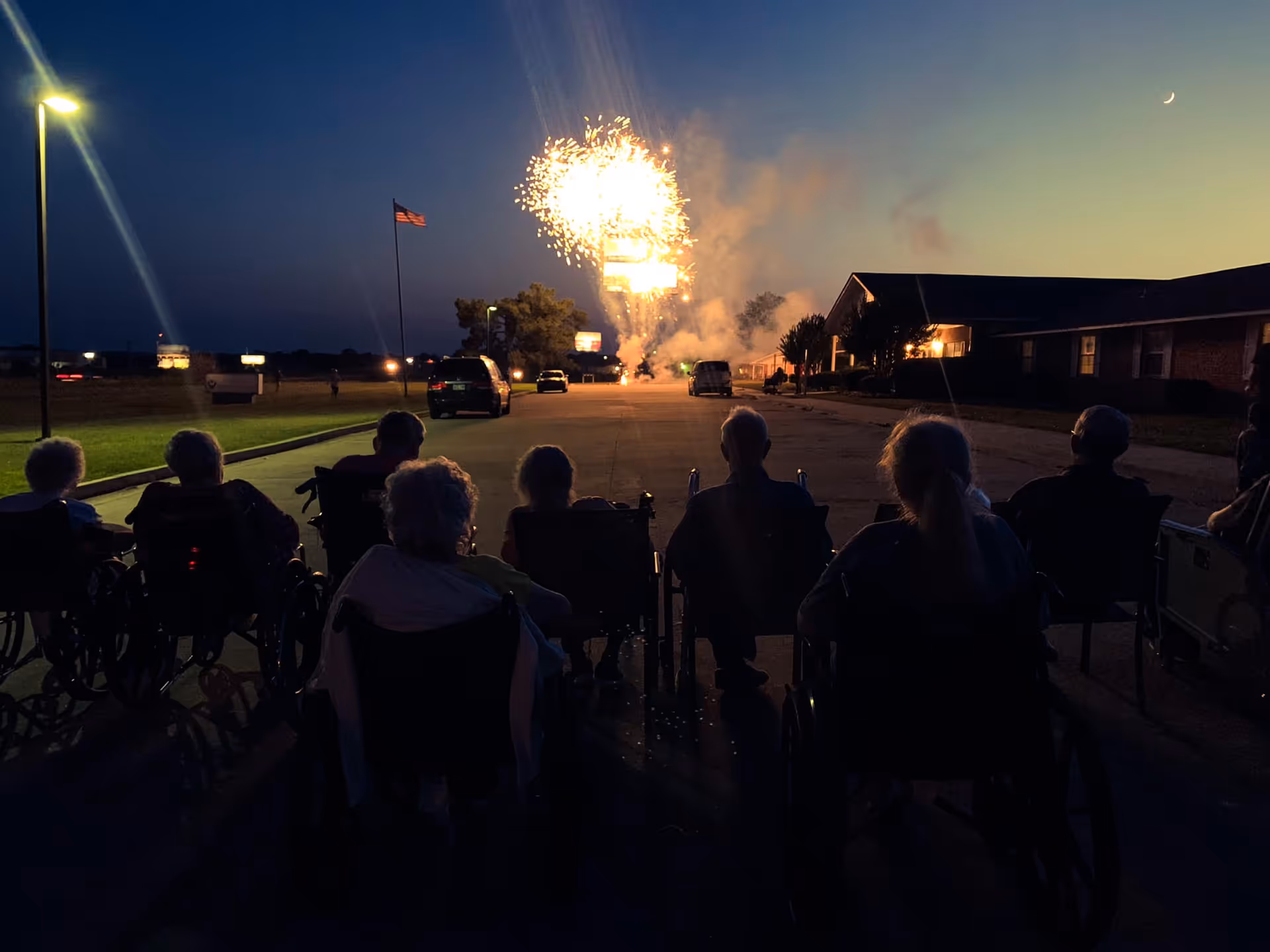 A group of elderly people, some in wheelchairs, sitting outside at dusk watching fireworks in the sky near a building with lights on.