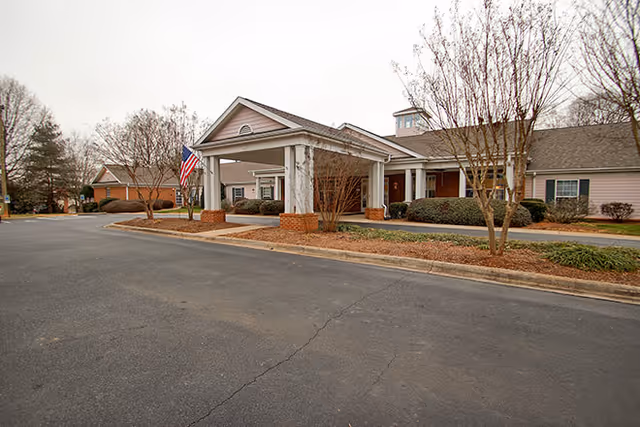 Exterior view of a single-story senior living facility building with a covered entrance supported by white columns and an American flag displayed near the entrance. The surrounding area includes a paved driveway, leafless trees, and some shrubs under an overcast sky.