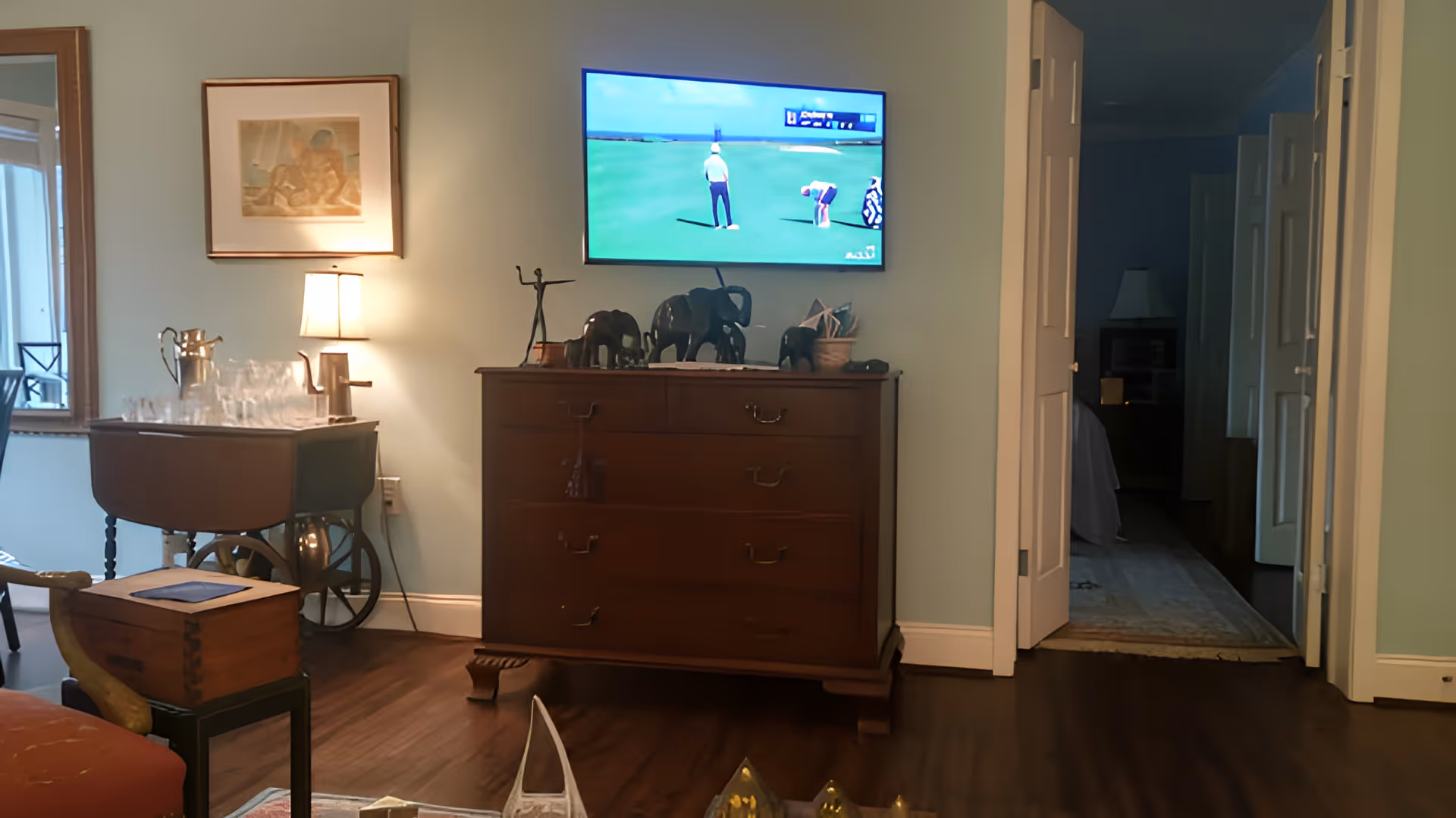 A cozy living room area with a wooden dresser topped with elephant figurines and a flat-screen TV mounted on the wall showing a golf game. To the left, there is a small bar cart with glassware and a lamp on the wall above it. A doorway leads to a dimly lit hallway with a glimpse of another room.