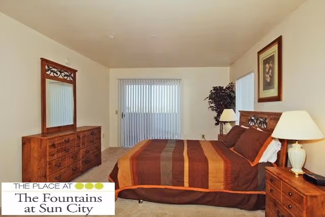 A bedroom with a large bed featuring a brown and orange striped bedspread, two bedside tables each with a lamp, a wooden dresser with a mirror, a potted plant in the corner, and vertical blinds covering a sliding glass door.