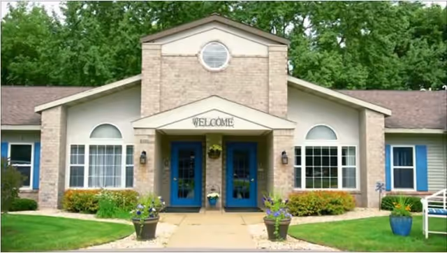 Front exterior view of a single-story building with beige brick and siding, featuring two blue entrance doors under a small gabled roof with a 'WELCOME' sign above. There are large windows on either side of the entrance, potted plants with flowers along the walkway, and green trees in the background.