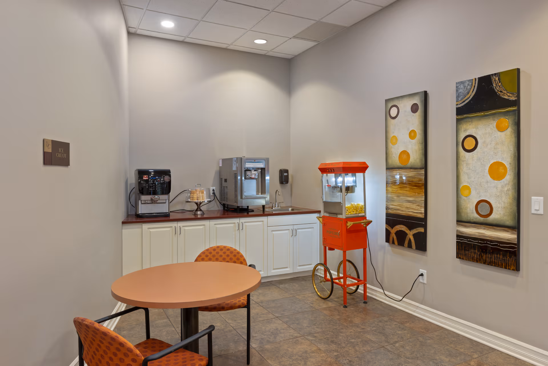 A small refreshment area in a senior living facility with a round table and two orange patterned chairs. On the countertop, there is a coffee machine, an ice cream machine, and a sink. A red popcorn machine is placed on the floor next to the counter. Two abstract paintings with circular designs hang on the wall.