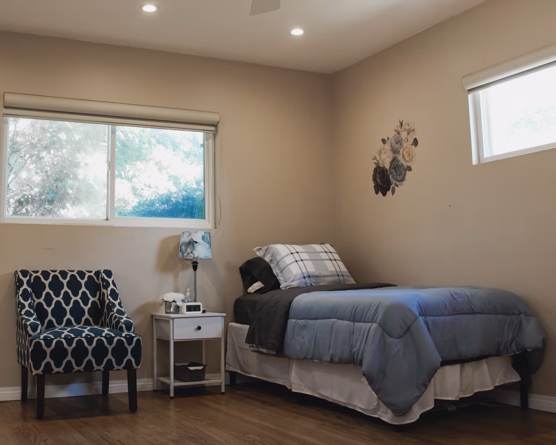 A simple bedroom with a single bed covered in gray and blue bedding, a white nightstand with a lamp and clock, a patterned armchair, two windows with blinds, and a floral wall decoration.