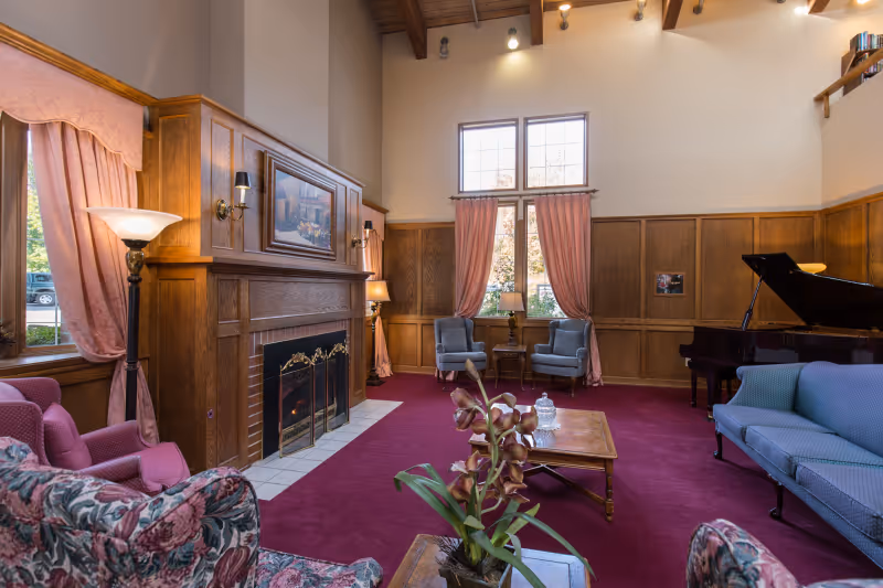 A spacious senior living facility common area with high ceilings, wood-paneled walls, and large windows with pink curtains. The room features a fireplace with a decorative screen, several upholstered chairs and sofas in floral and solid patterns, a wooden coffee table with a plant and decorative items, and a grand piano in the corner.