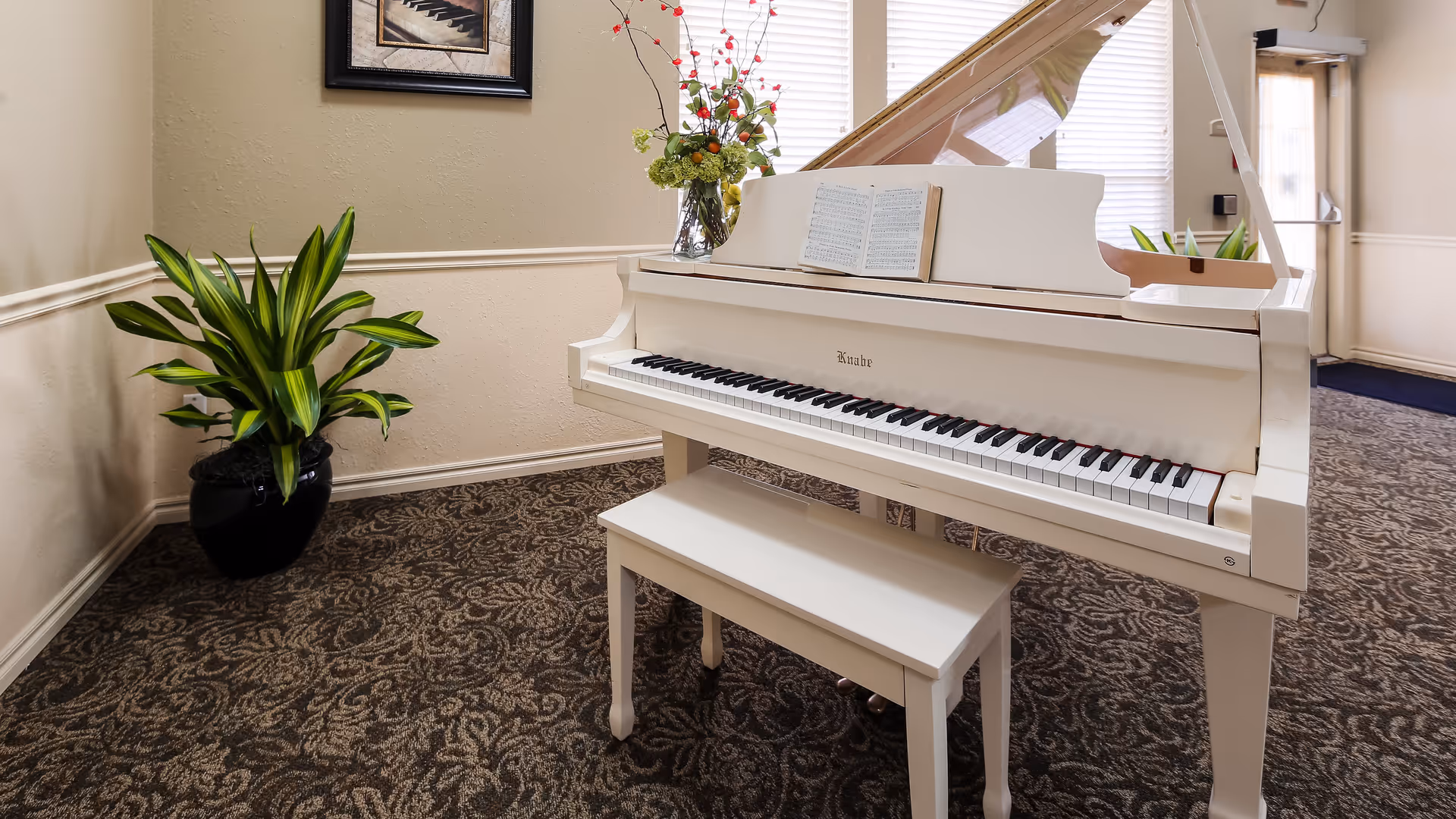 A white grand piano with an open lid and sheet music on the stand, placed on a patterned carpet in a well-lit room. A matching white bench is in front of the piano. There is a potted green plant to the left and a vase with flowers on top of the piano. A framed picture hangs on the beige wall behind the piano, and a door with a window is visible in the background.