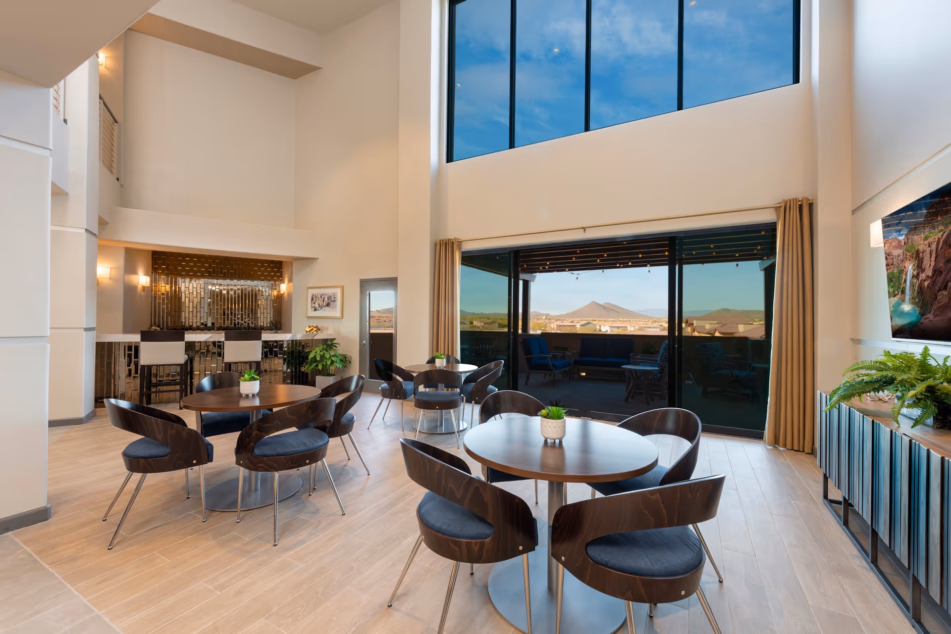 A spacious and modern common area with round tables and chairs arranged for seating. Large floor-to-ceiling windows provide a view of an outdoor patio and distant mountains. The room features light wood flooring, a bar area with stools, and decorative plants. The high ceiling and natural light create an open and inviting atmosphere.