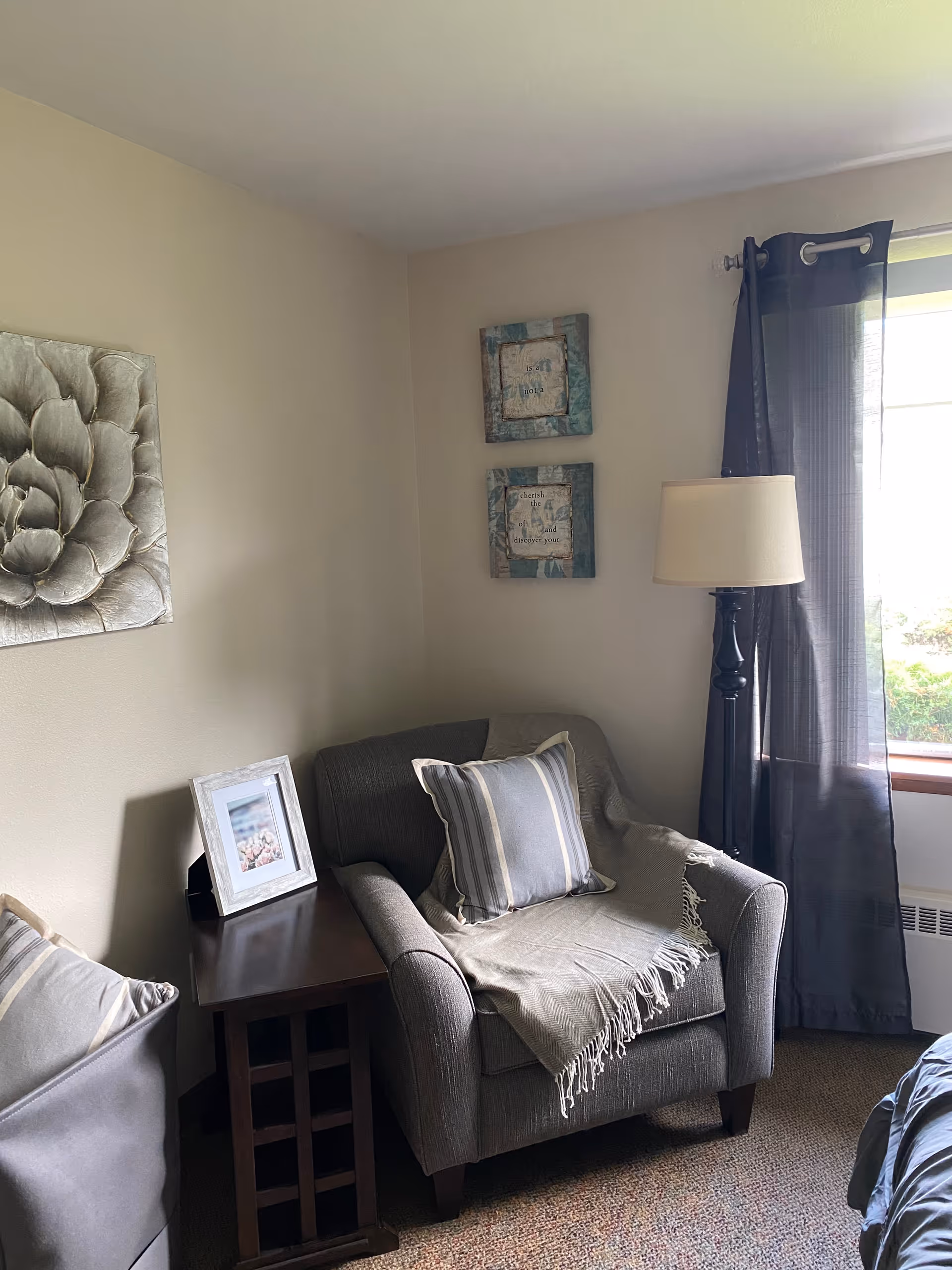 A cozy corner of a room featuring a gray upholstered armchair with a striped pillow and a light throw blanket. Next to the chair is a dark wooden side table with a framed photo. On the wall above the chair are two decorative framed quotes, and to the left is a large floral wall art piece. A window with sheer dark curtains allows natural light into the room.