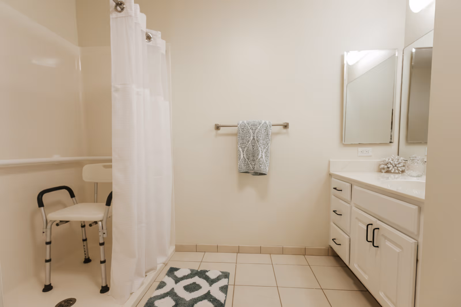 A clean, accessible bathroom with a white shower curtain, a shower chair with black handles inside the shower, a towel hanging on a rack on the wall, a white vanity with drawers and cabinets, a large mirror above the sink, and a patterned bath mat on the tiled floor.