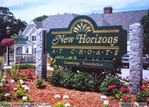 Outdoor view of the New Horizons At Choate facility sign surrounded by a landscaped garden with flowers and shrubs, a gazebo, and a building in the background under a blue sky.