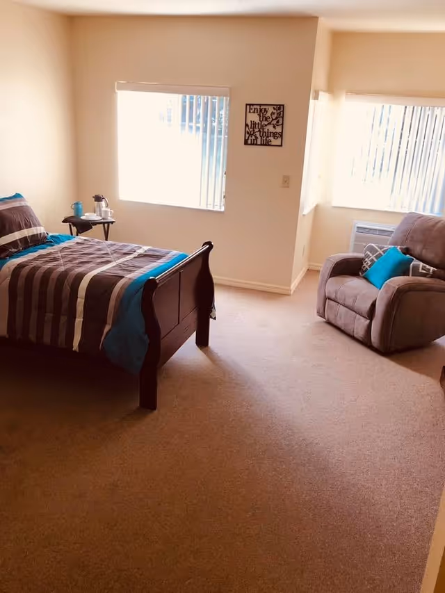 A bedroom with a wooden bed featuring striped bedding in shades of brown, beige, and blue. Next to the bed is a small table with a teapot and cup. There is a comfortable armchair with a blue pillow near two windows with vertical blinds. A wall decoration with the phrase 'Enjoy the little things in life' is visible between the windows.
