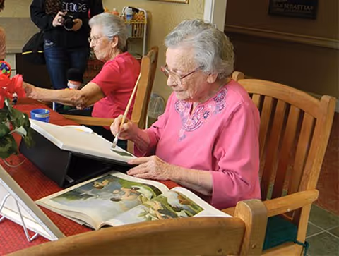 Two elderly women sitting at a wooden table engaged in painting and looking at an art book. One woman in a pink top is painting with a brush, while the other woman in a red top is also involved in an activity. A person in the background is taking a photo.