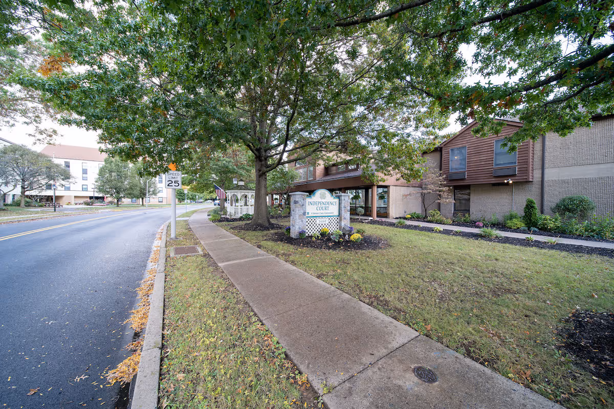 Sidewalk leading to the entrance of Independence Court of Quakertown, a senior living facility. The building is surrounded by green grass, trees, and landscaped areas. A speed limit sign of 25 mph is visible along the adjacent road.