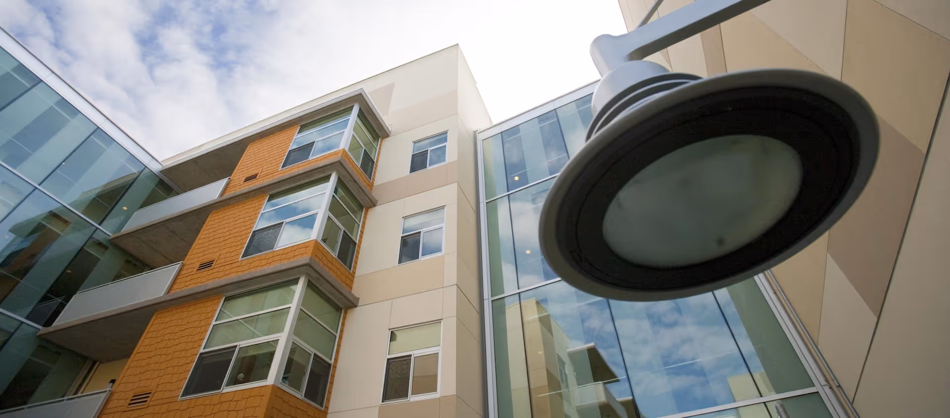 Upward view of a modern multi-story building with large windows and a mix of beige and orange exterior walls under a partly cloudy sky. A large outdoor light fixture is prominently visible in the foreground.
