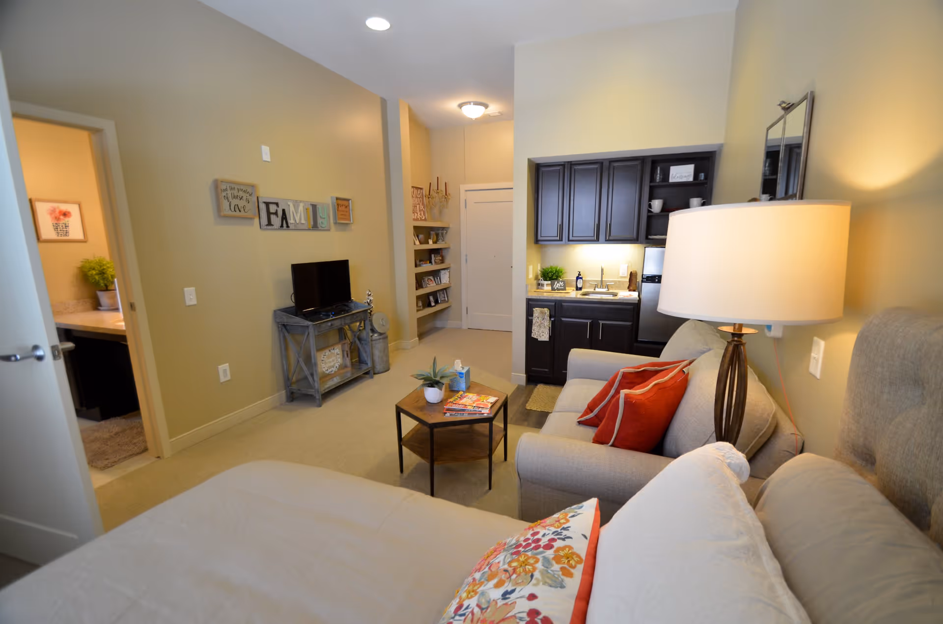 Interior view of a cozy senior living facility room at The Goldton at Jones Farm featuring a bed with floral and white pillows in the foreground, a small sitting area with a gray sofa and red pillows, a wooden coffee table with magazines and a plant, a lamp on a side table, a small kitchenette with dark cabinets and a sink, and a TV on a stand against a beige wall decorated with family-themed wall art. A doorway leads to a bathroom with a plant on the counter.