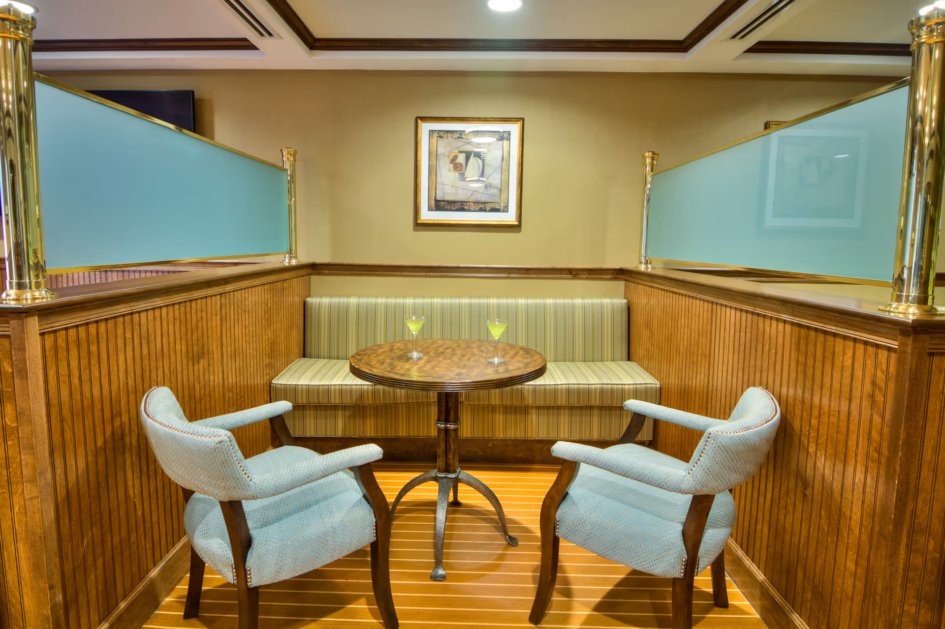 A cozy seating area with two light blue upholstered armchairs and a round wooden table in between. Behind the table is a built-in bench with striped upholstery. The walls are wood-paneled with frosted glass dividers on top, and a framed abstract painting hangs on the beige wall above the bench.