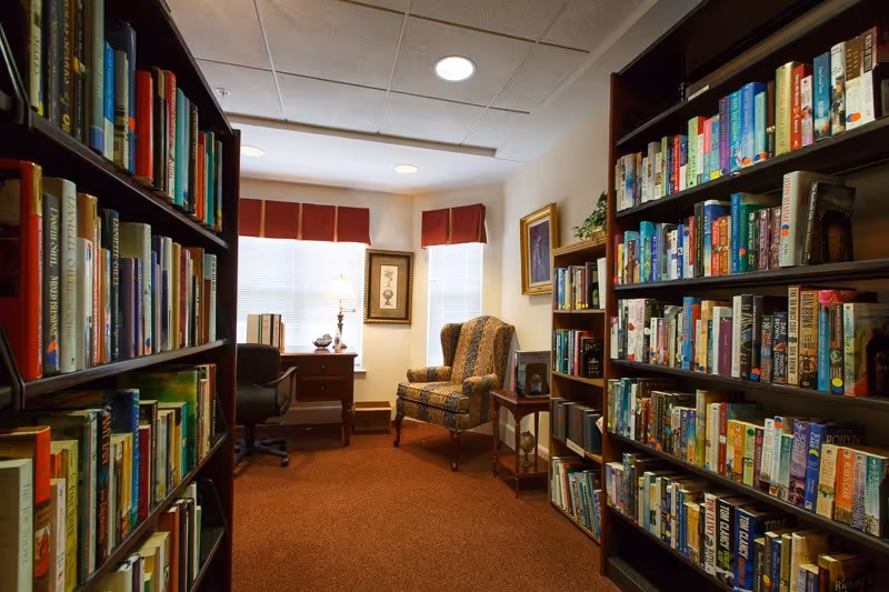 Small library/reading room with bookshelves on both sides, an upholstered armchair, desk, and lamp by a window.