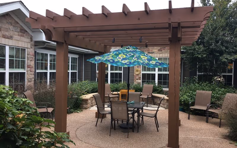 Outdoor patio area with a wooden pergola overhead, a glass table with four chairs, and a colorful blue and green patterned umbrella. Surrounding the patio are plants, bushes, and a stone building with multiple windows.