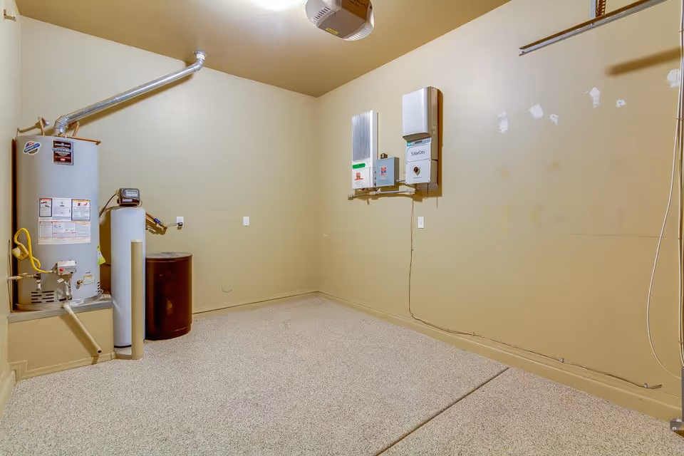 Utility room with a water heater, tanks and wall-mounted electrical units against beige walls and a speckled floor.