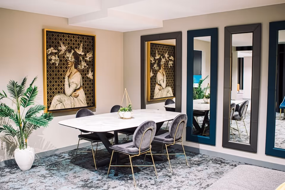 A stylish indoor dining area with a white marble table surrounded by gray velvet chairs, large framed artwork, tall mirrors, and a potted plant.