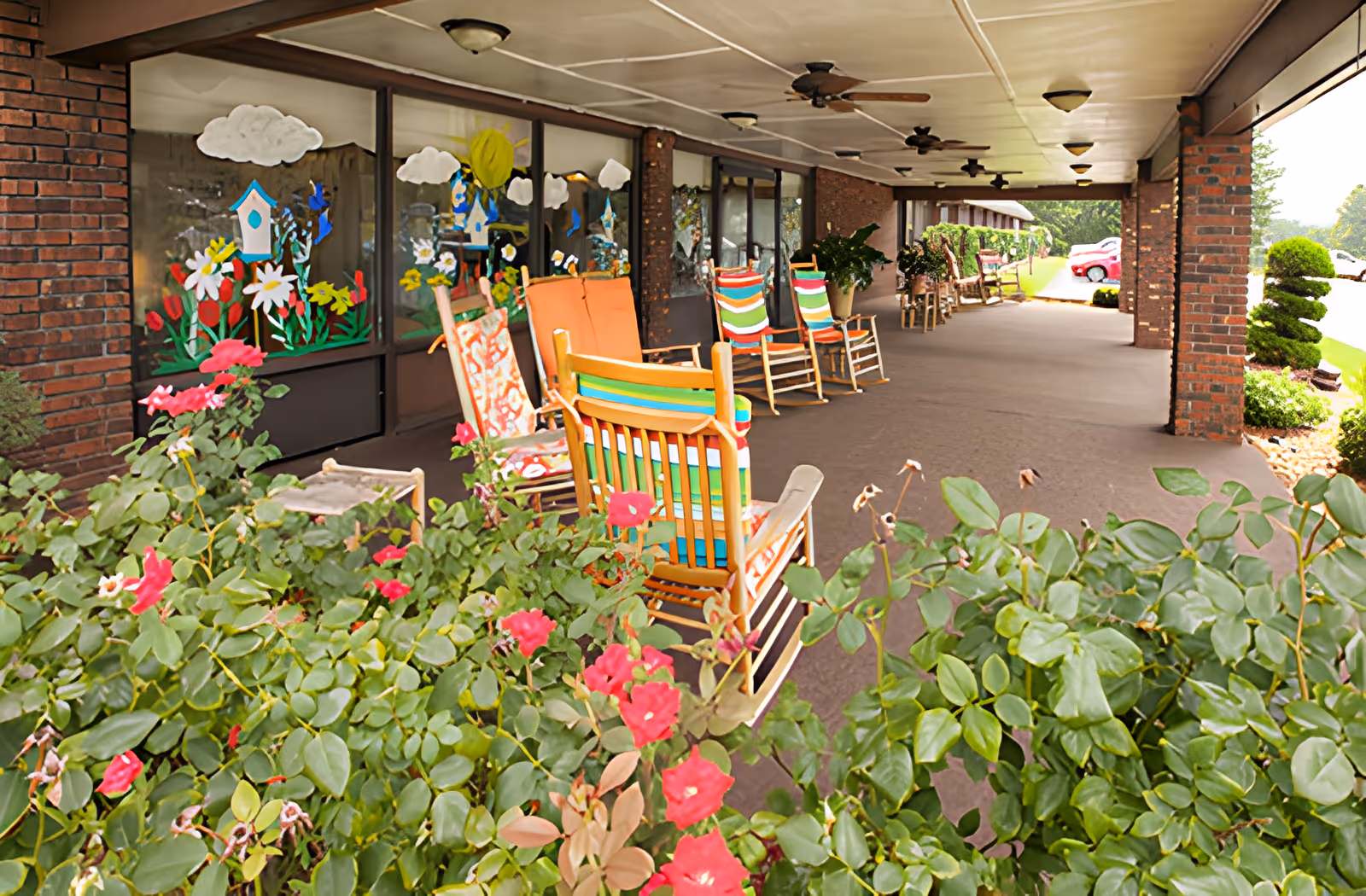 Covered outdoor patio area with several colorful rocking chairs arranged along the wall of a brick building. The windows are decorated with painted flowers, clouds, and birdhouses. In the foreground, there are green bushes with pink flowers. Ceiling fans are mounted on the patio ceiling, and a parking lot with cars is visible in the background.