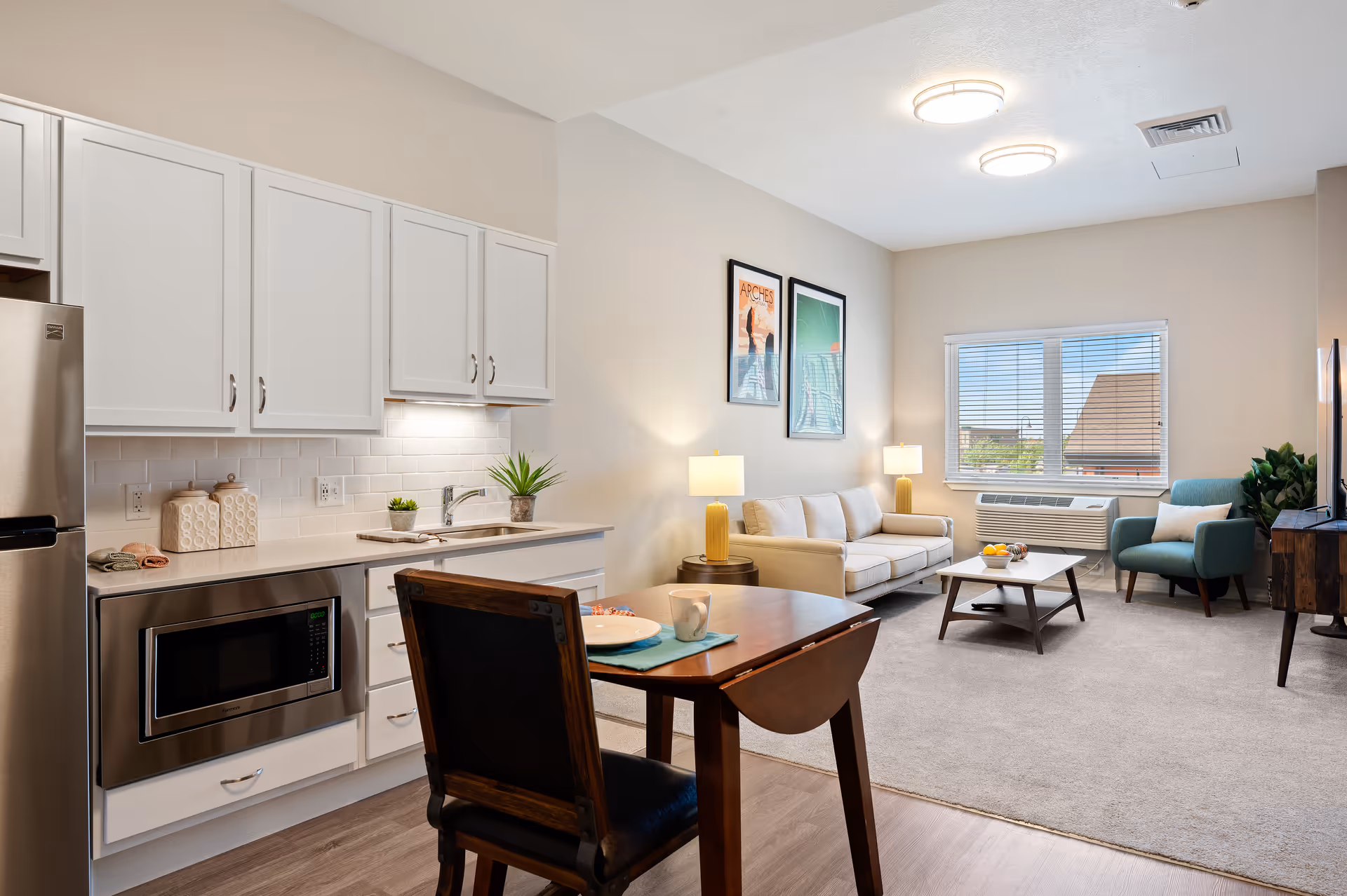 A bright and modern assisted living apartment featuring a small kitchen area with white cabinets, a stainless steel microwave, and a sink. Adjacent to the kitchen is a wooden dining table with a single chair, set with a plate and mug. The living area includes a beige sofa, a blue armchair, a coffee table with a bowl of fruit, two yellow table lamps, and framed artwork on the wall. A window with blinds lets in natural light, and a TV is positioned on a wooden stand.