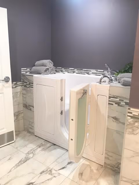 A modern walk-in bathtub with an open door in a bathroom featuring marble-patterned floor and wall tiles, a gray painted wall, and neatly folded gray towels placed on the tub edge.