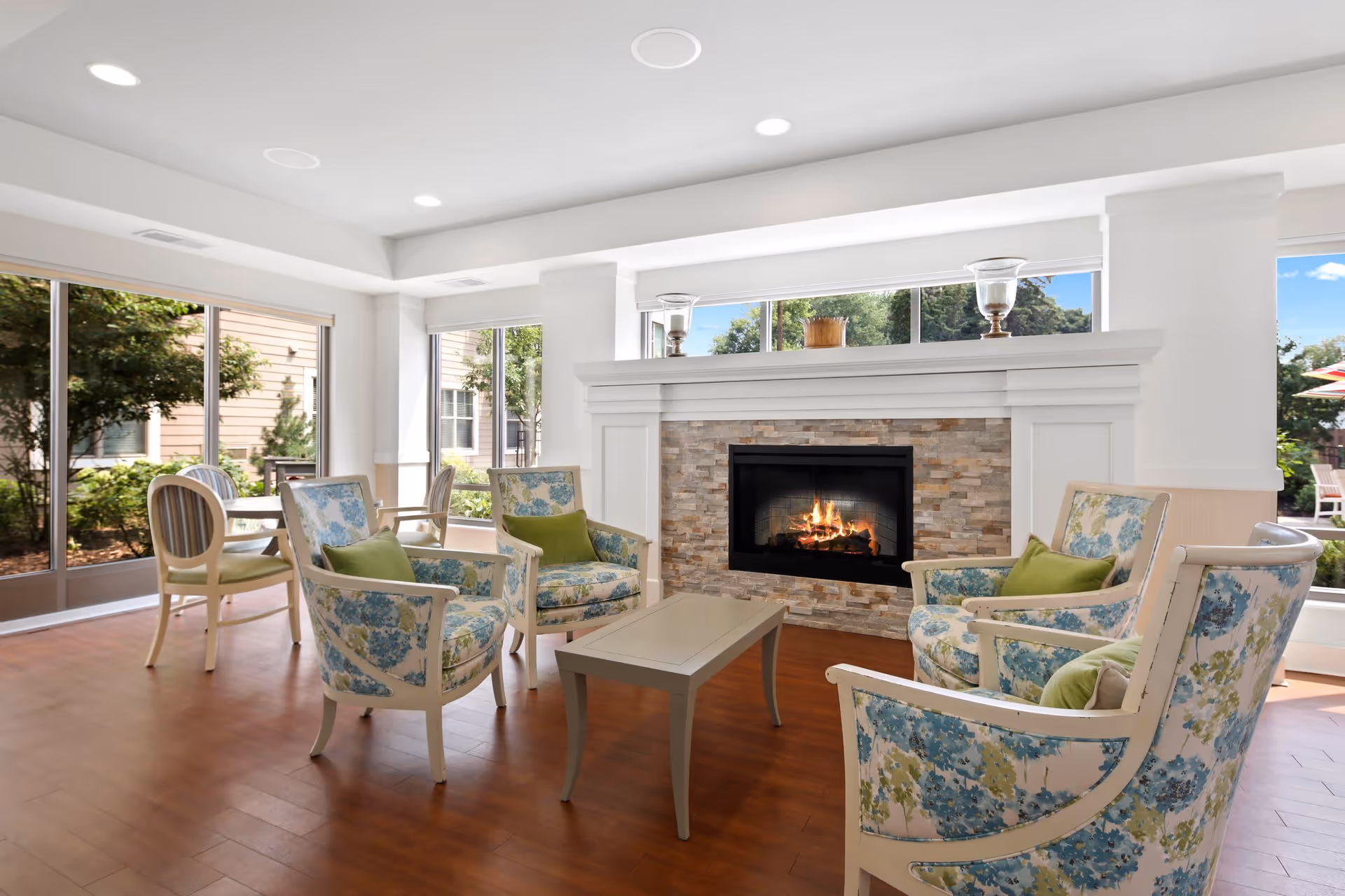 A bright and cozy sitting area with four floral upholstered armchairs arranged around a small rectangular coffee table in front of a stone fireplace with a fire burning. Large windows surround the room, letting in natural light and offering views of greenery outside. The floor is wooden, and the ceiling has recessed lighting.