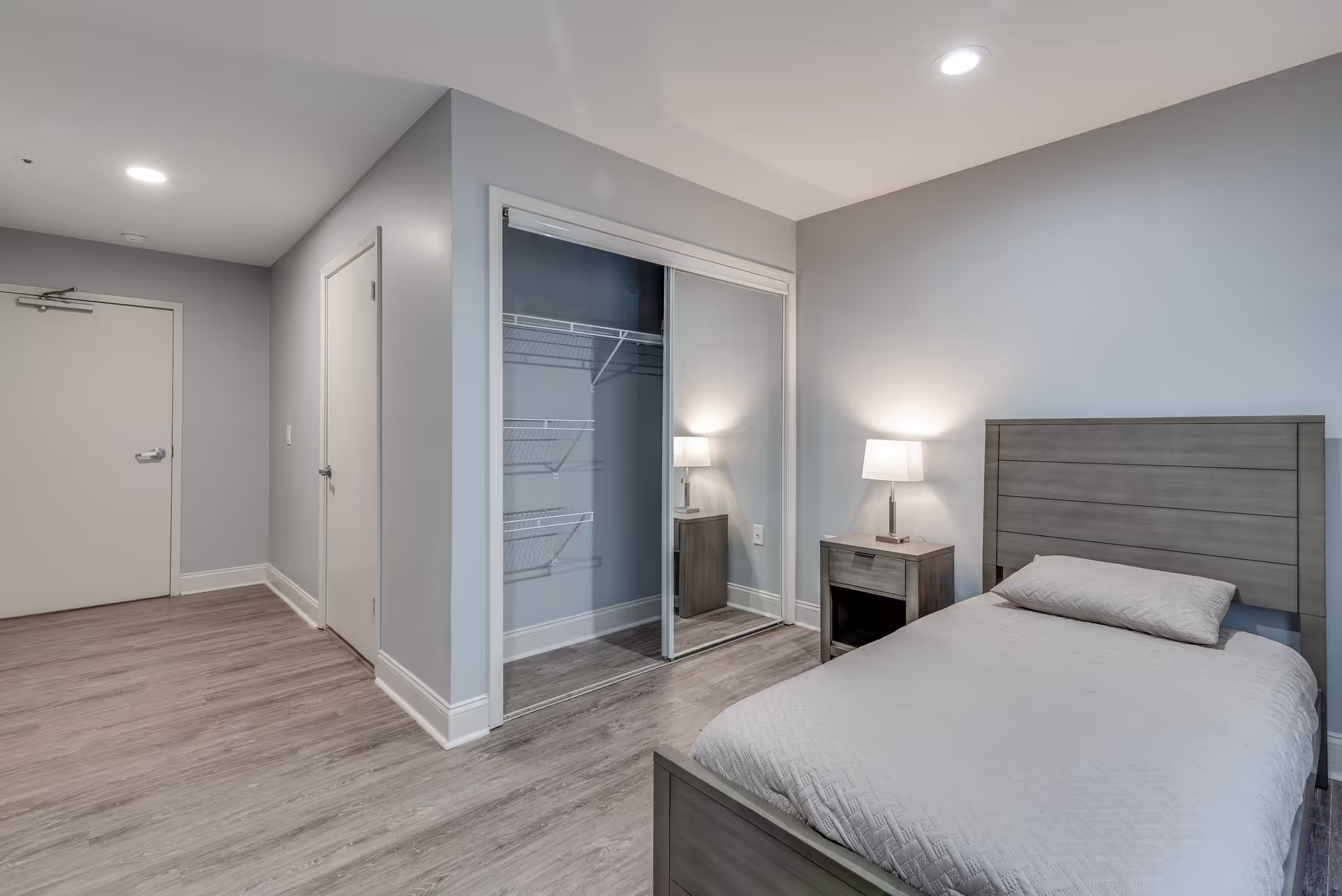 A simple bedroom with a single bed featuring a gray wooden headboard and a light gray bedspread. Next to the bed is a small wooden nightstand with a lamp on it. The room has light gray walls and wood flooring. There is a closet with sliding mirrored doors and wire shelving inside. Two closed doors are visible on the left side of the room.