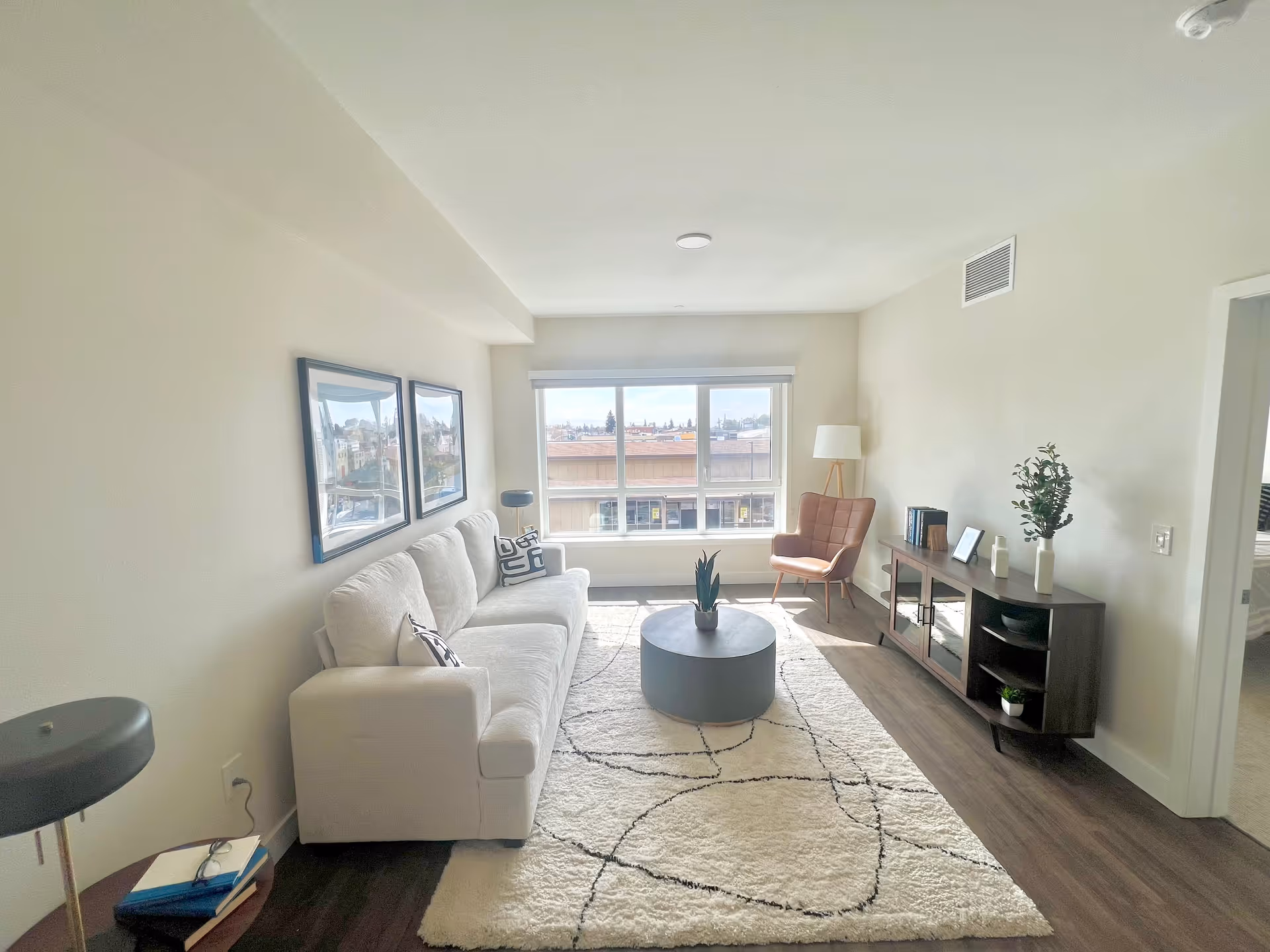 A bright living room with a large window letting in natural light. The room features a white sofa with patterned pillows, a round coffee table with a small plant, a modern armchair, a floor lamp, and a wooden media console with decorative items. The floor is wood with a light-colored area rug, and two framed pictures hang on the wall above the sofa.