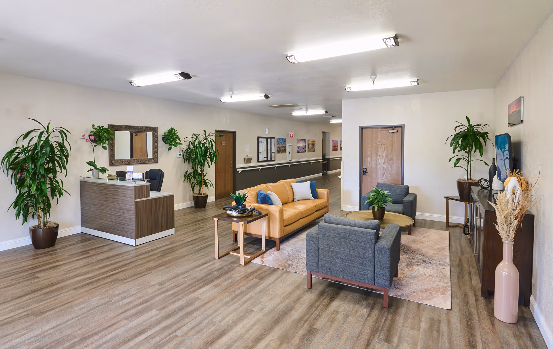 Bright reception lobby with a desk to the left and a seating area of a tan sofa and gray chairs on a rug, surrounded by potted plants.