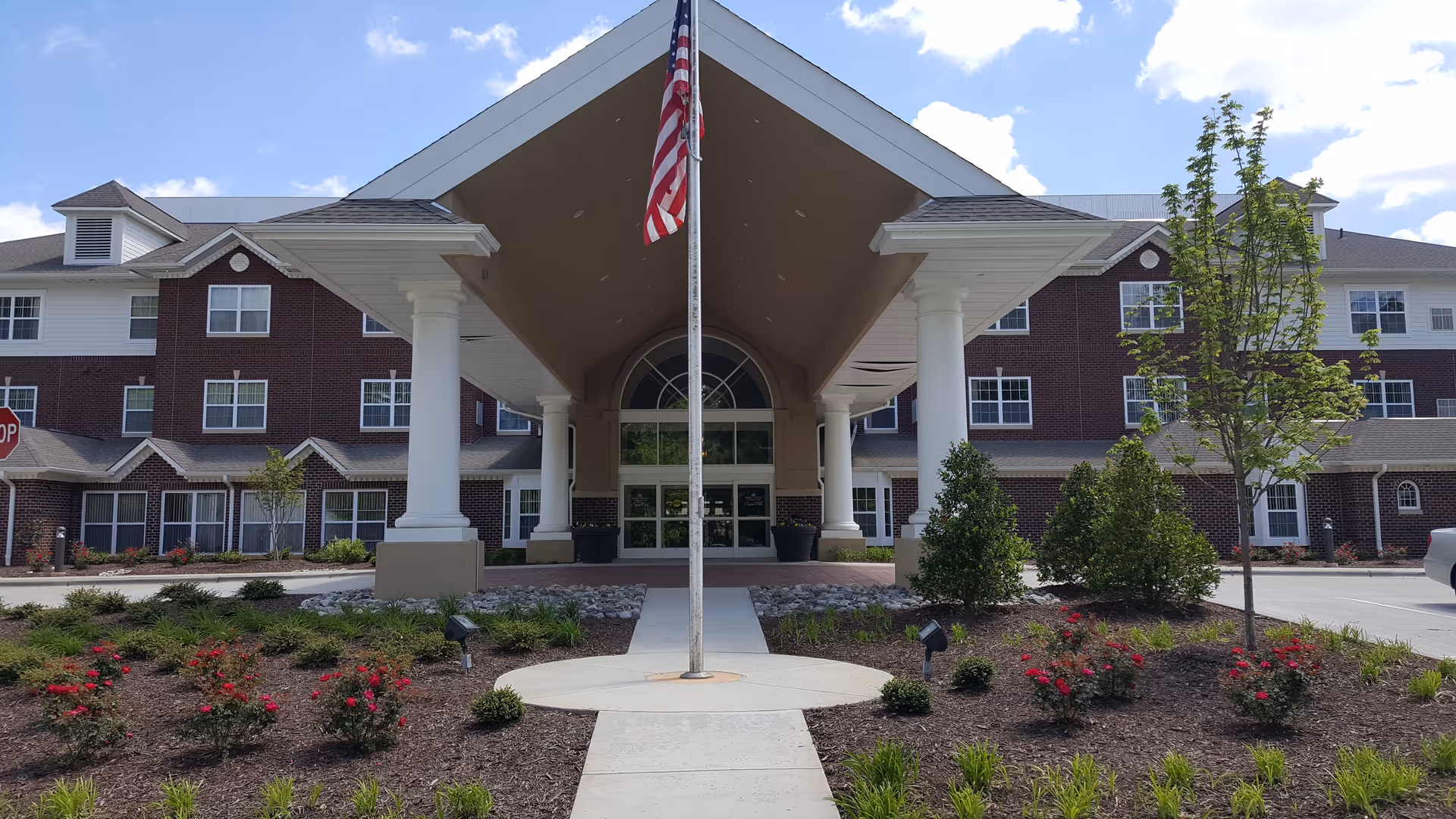 Front exterior view of Capital Oaks Retirement Resort featuring a large covered entrance with white columns, an American flag on a flagpole in the center, landscaped flower beds with red flowers and greenery, and a multi-story brick building with many windows under a partly cloudy sky.