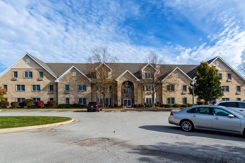 Front exterior view of a large, three-story brick building with multiple windows and a central entrance under an archway. There are a few cars parked in the driveway in front of the building, and some trees and shrubs around the entrance. The sky is partly cloudy.