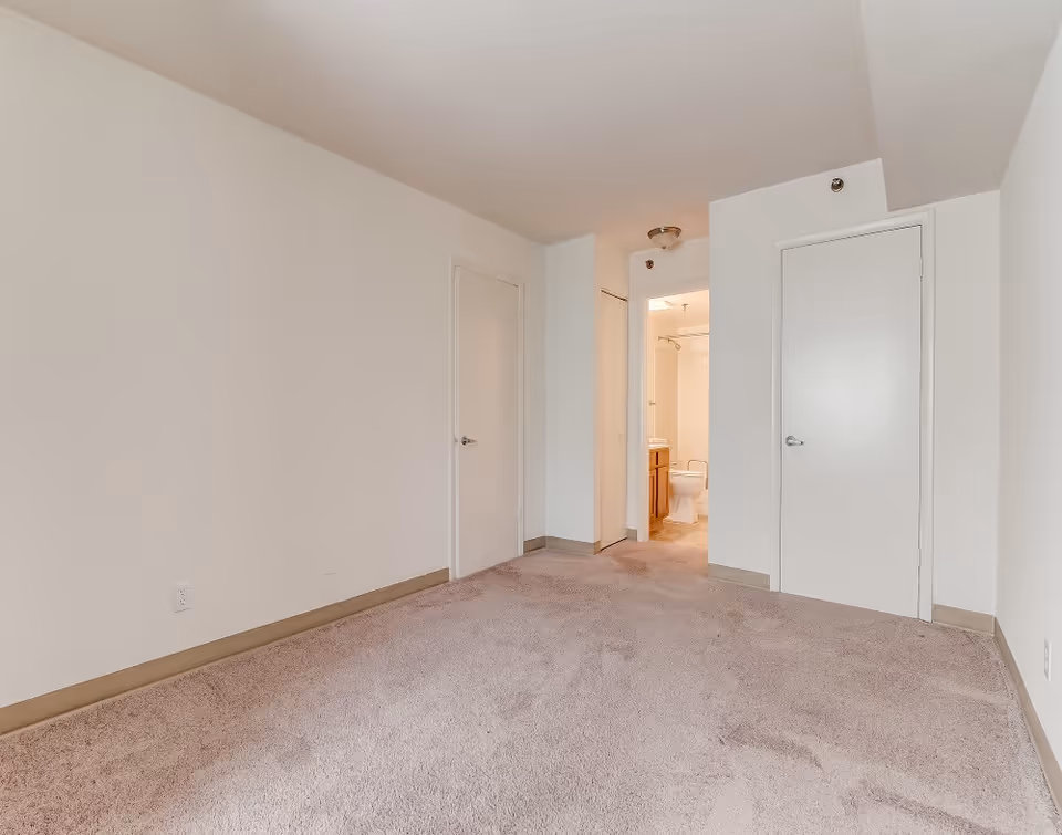 Empty room with beige carpet and white walls featuring three closed doors and one open door leading to a bathroom with a toilet and vanity visible.