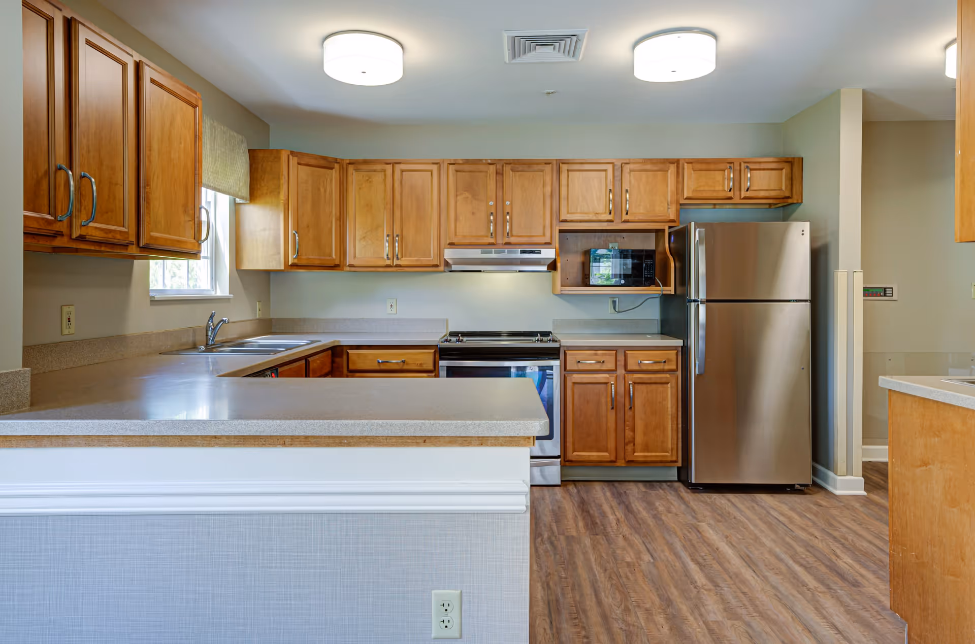 A clean and modern kitchen with wooden cabinets, a stainless steel refrigerator, a microwave, an oven with a stove, and a double sink under a window. The kitchen has light-colored countertops and wood-style flooring, with two ceiling lights providing illumination.