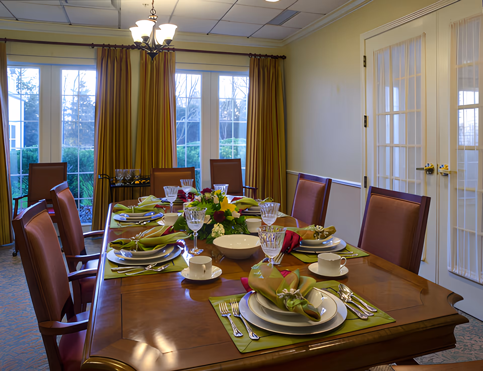 A formal dining room with a set wooden table, place settings and chairs, chandelier, and large windows with curtains.