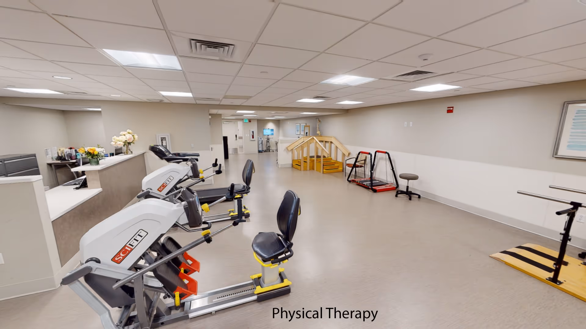 A spacious physical therapy room with exercise equipment including SciFit machines, a small set of wooden stairs with handrails, and parallel bars for walking exercises. The room has a light-colored floor and walls, a drop ceiling with fluorescent lighting, and a reception desk with flowers on it.