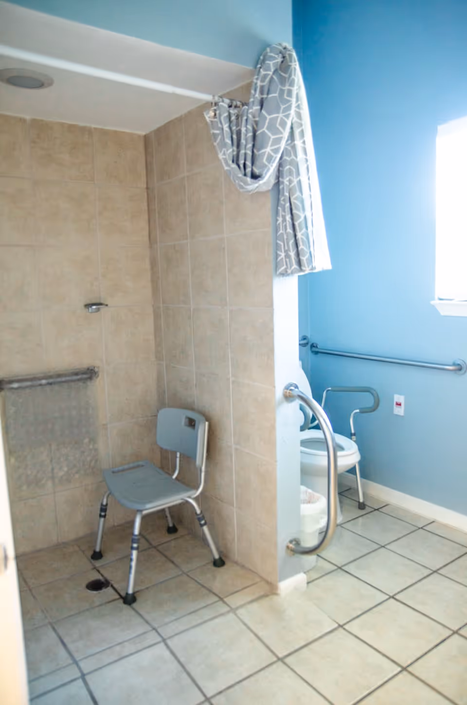 A bathroom with beige tiled walls and floor, featuring a shower area with a gray shower chair and a grab bar. Adjacent to the shower is a toilet with grab bars on both sides, a blue wall, and a window letting in natural light. A patterned shower curtain is pulled to the side.