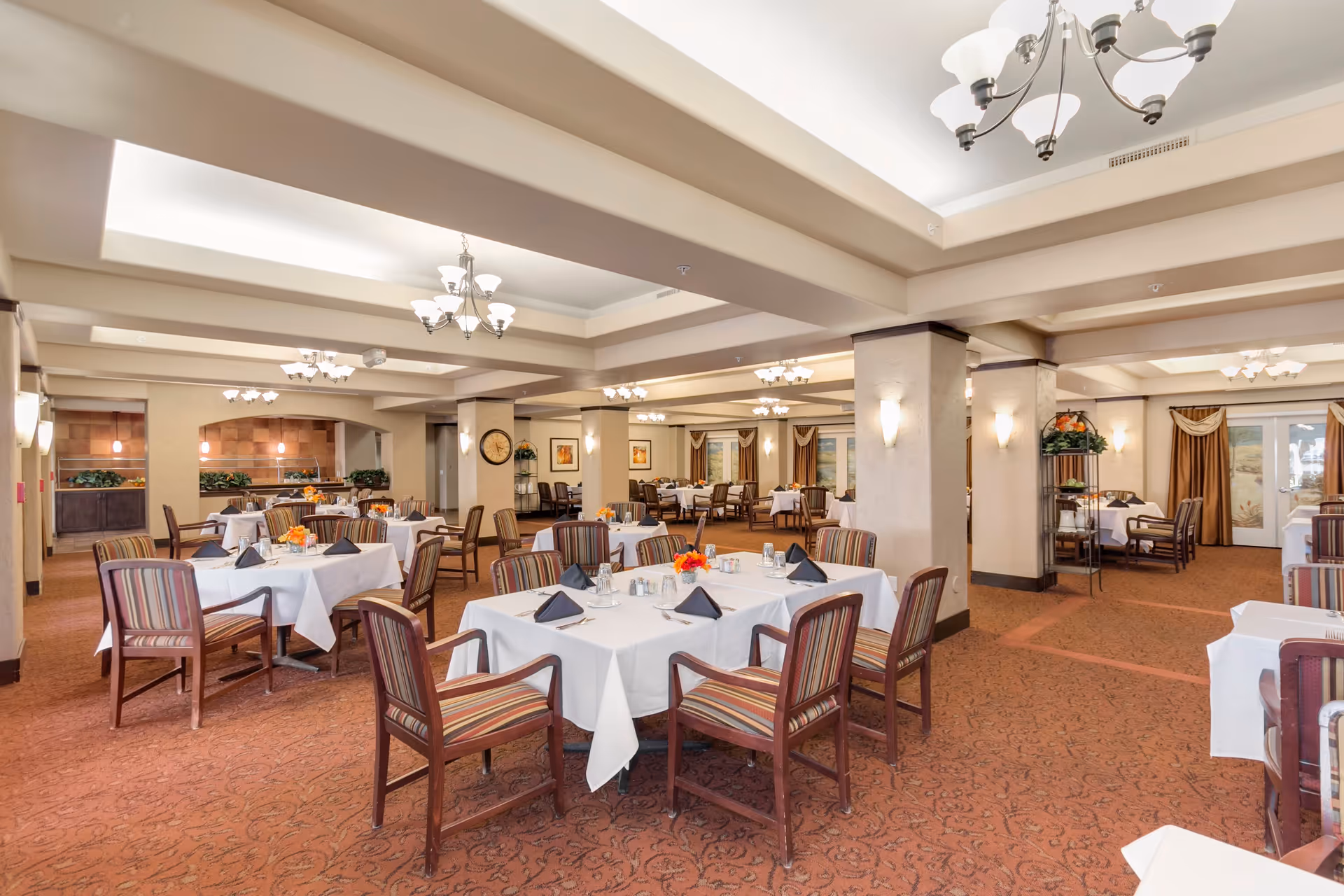 A spacious dining room with multiple tables covered in white tablecloths, each set with black napkins, glassware, and small floral centerpieces. The room features carpeted floors, beige walls with pillars, and several chandeliers hanging from the ceiling providing warm lighting. Chairs with striped upholstery surround the tables, and there are decorative plants and framed artwork on the walls.