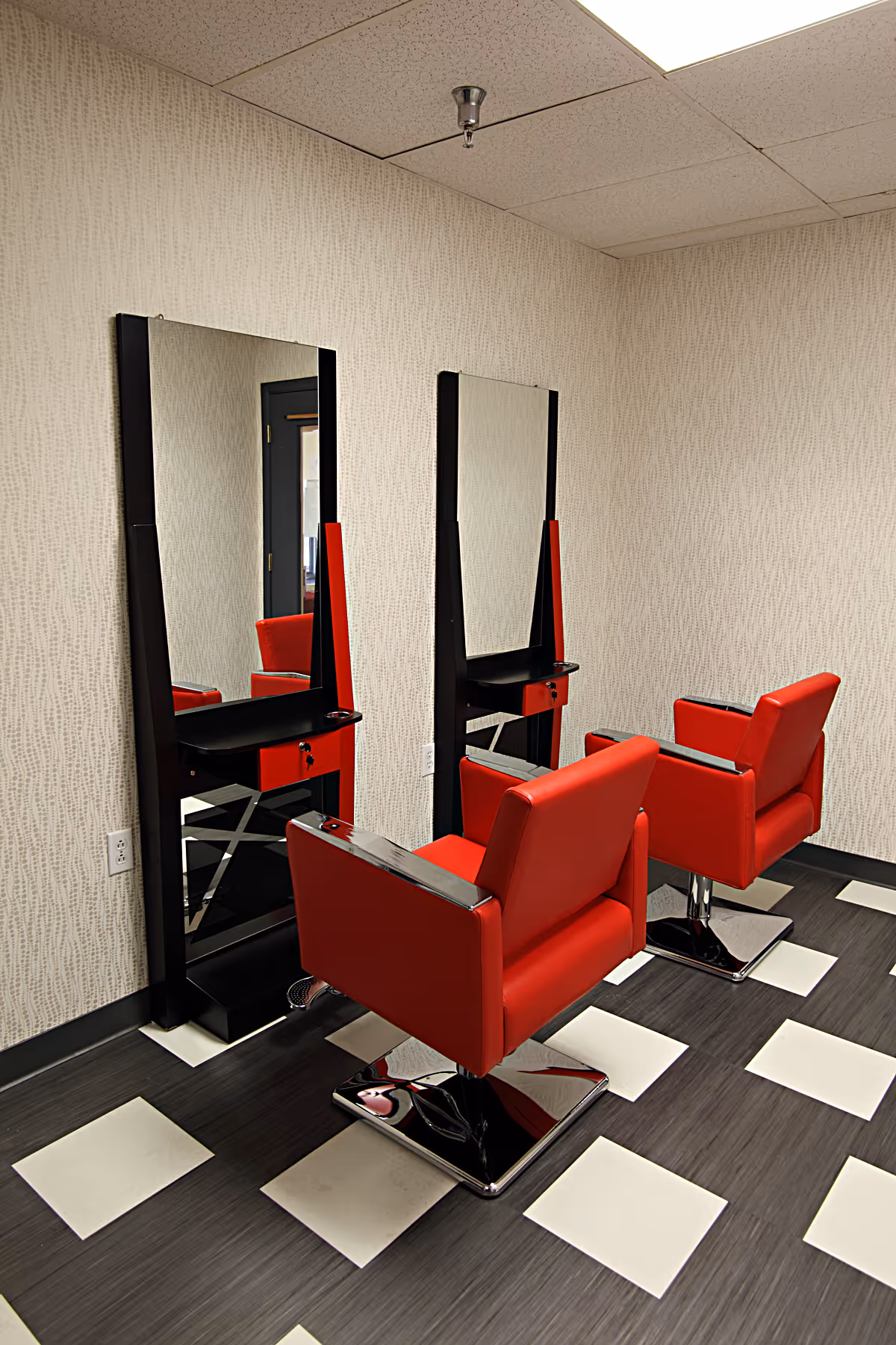 Two red salon chairs at styling stations with mirrors in a small hair salon area with a black-and-white tiled floor.
