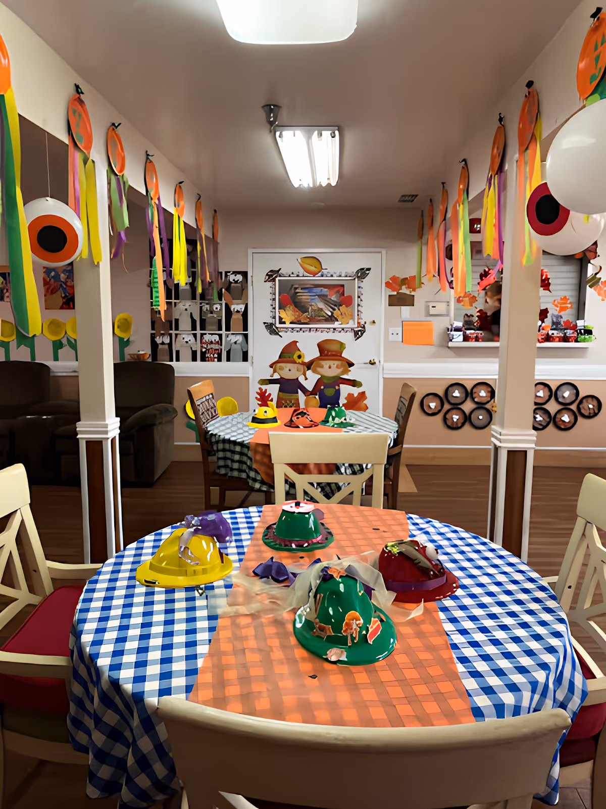 A decorated dining area with tables covered in blue and white checkered tablecloths and orange runners. The tables have colorful craft hats on them. The room is adorned with hanging orange and yellow paper decorations and large eyeball balloons. In the background, there are two scarecrow figures on a door and various wall decorations.