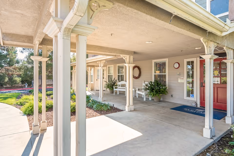 Covered front porch entrance of a senior living facility with white columns, benches, potted plants and a red door.