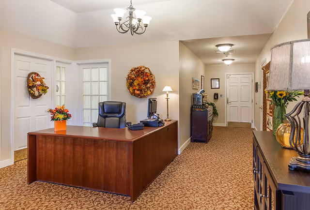 A reception area with a wooden desk and office chair, decorative wreaths on the walls, and a carpeted hallway leading to doors.