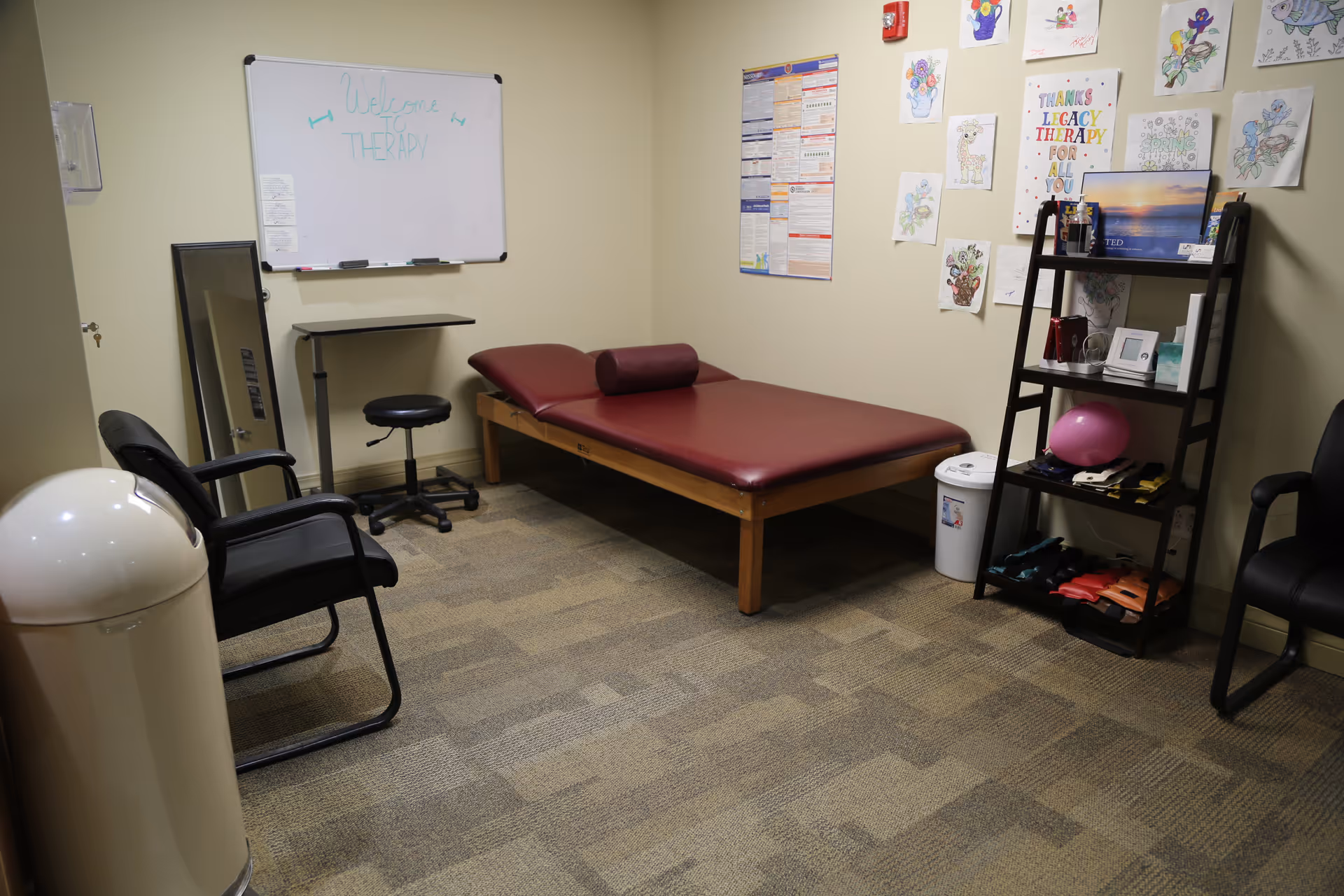 A therapy room with a maroon padded therapy table, two black chairs, a whiteboard with 'Welcome to Therapy' written on it, a small adjustable table, a full-length mirror, and a shelving unit holding various therapy tools and colorful drawings on the wall.