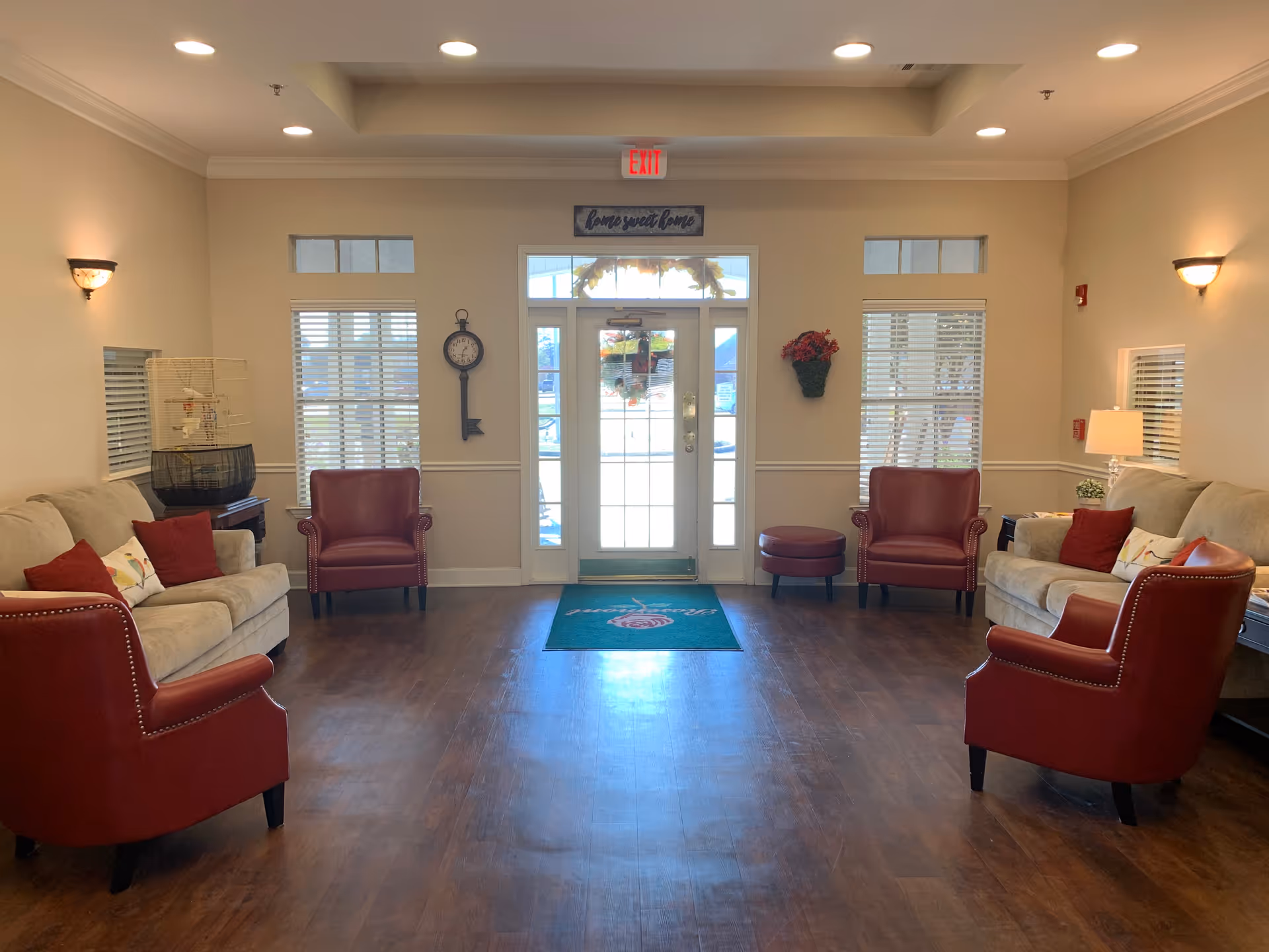 A cozy assisted living facility lobby with two beige sofas and four red armchairs arranged around a wooden floor. The entrance door is centered with windows on either side, and a 'home sweet home' sign above the door. There is a clock on the left wall and a wall-mounted flower basket on the right. The room is softly lit with wall sconces and a table lamp.