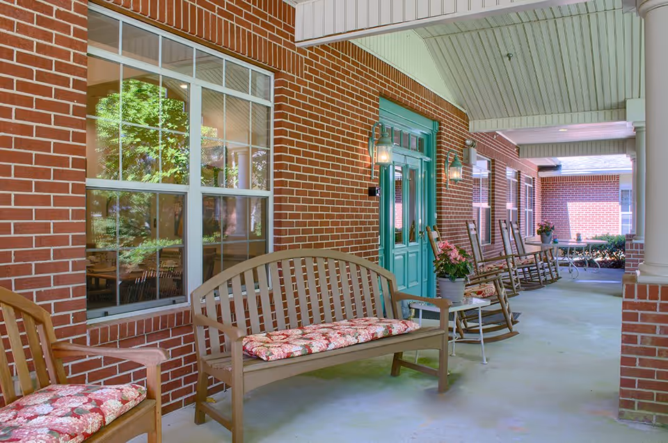 Covered outdoor patio area with brick walls and teal double doors. The patio is furnished with wooden benches and rocking chairs, all with floral cushions. There are potted plants on small tables and wall-mounted lantern-style lights beside the doors and windows.