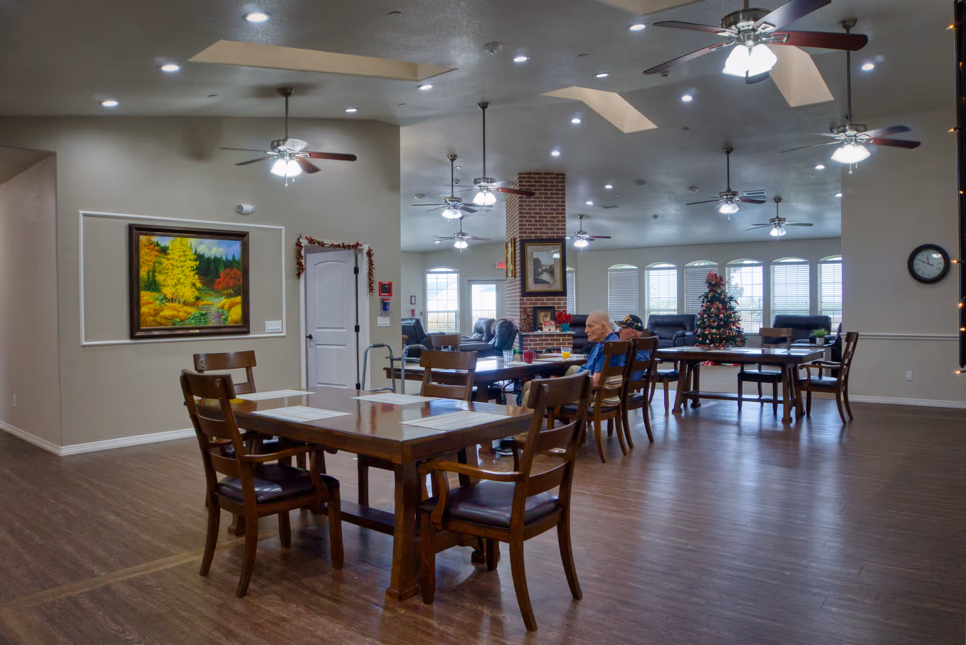A spacious common area in an assisted living facility with wooden tables and chairs arranged for dining or socializing. Ceiling fans with lights hang from the ceiling. There is a brick fireplace in the center with framed artwork above it. Large windows with blinds let in natural light. A decorated Christmas tree is visible near the windows. Two elderly individuals are seated at one of the tables.