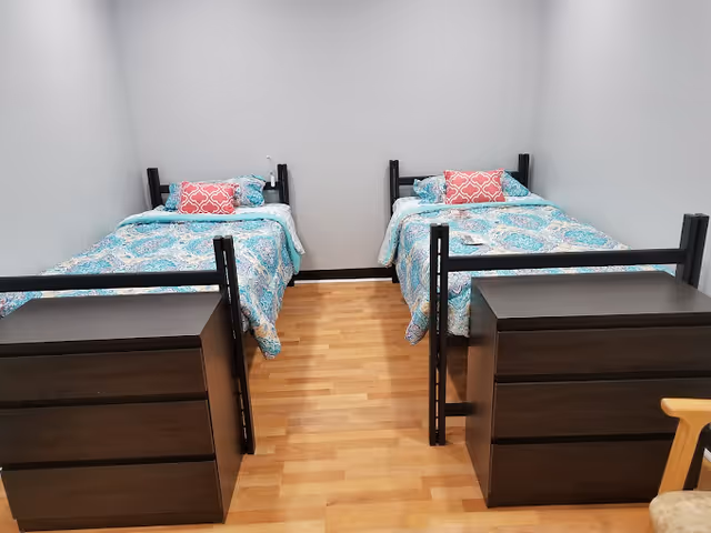 A simple bedroom with two twin beds side by side, colorful patterned bedding, and dark dressers at the foot of each bed.
