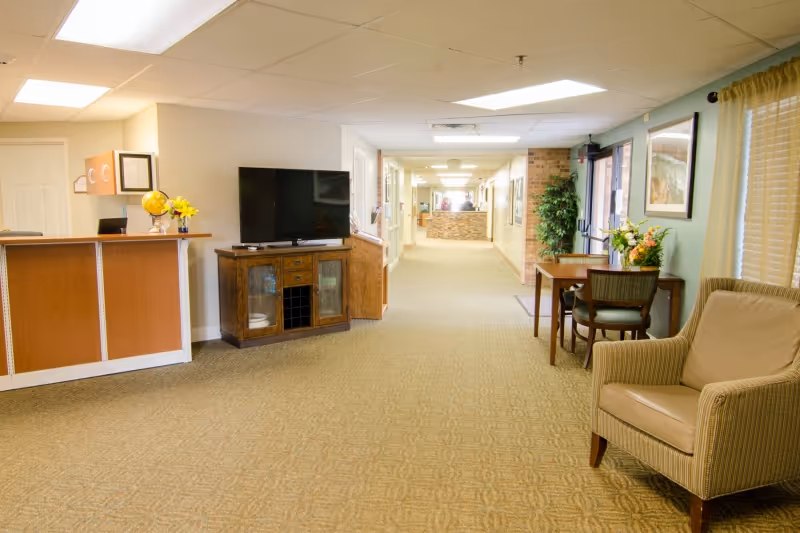 Bright common area of a senior living facility with a reception desk, a television on a wooden cabinet, chairs and a long hallway.