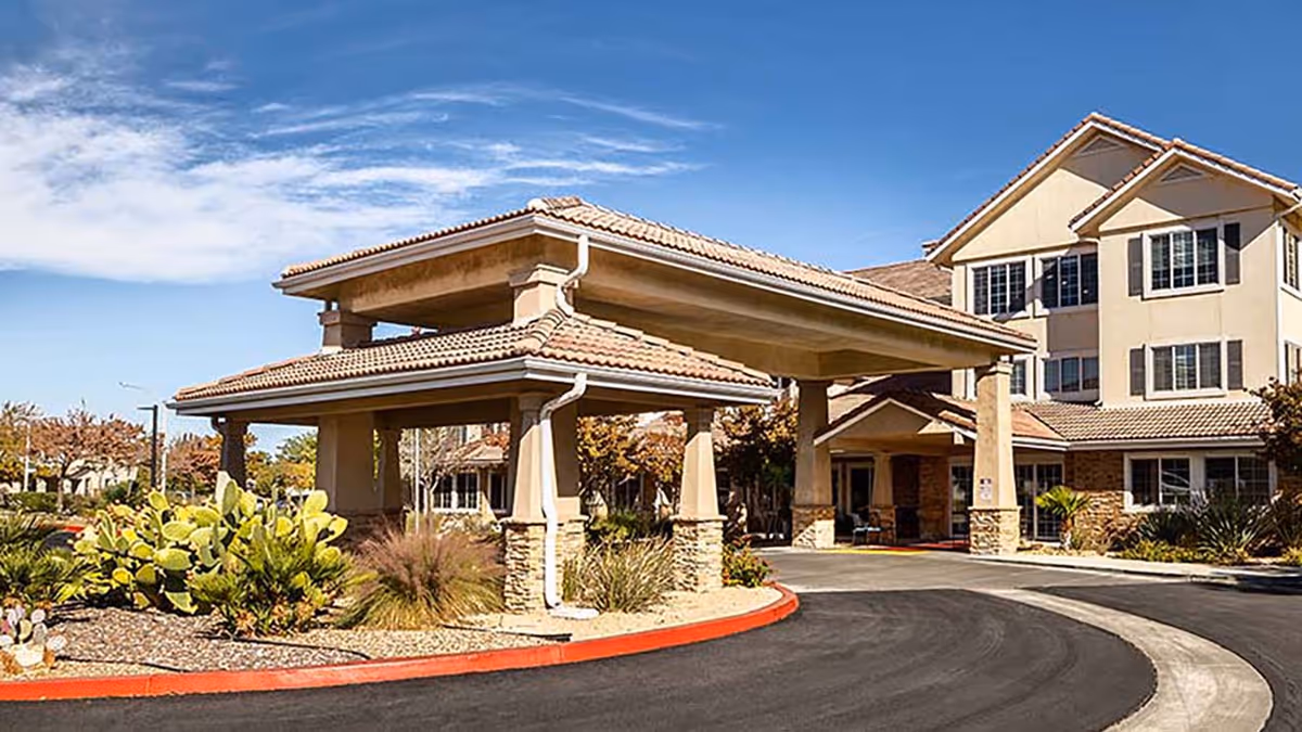 Exterior view of Holiday Rancho Village senior living facility showing the main entrance with a covered driveway, beige building with multiple windows, and desert landscaping including cacti and shrubs under a blue sky with some clouds.