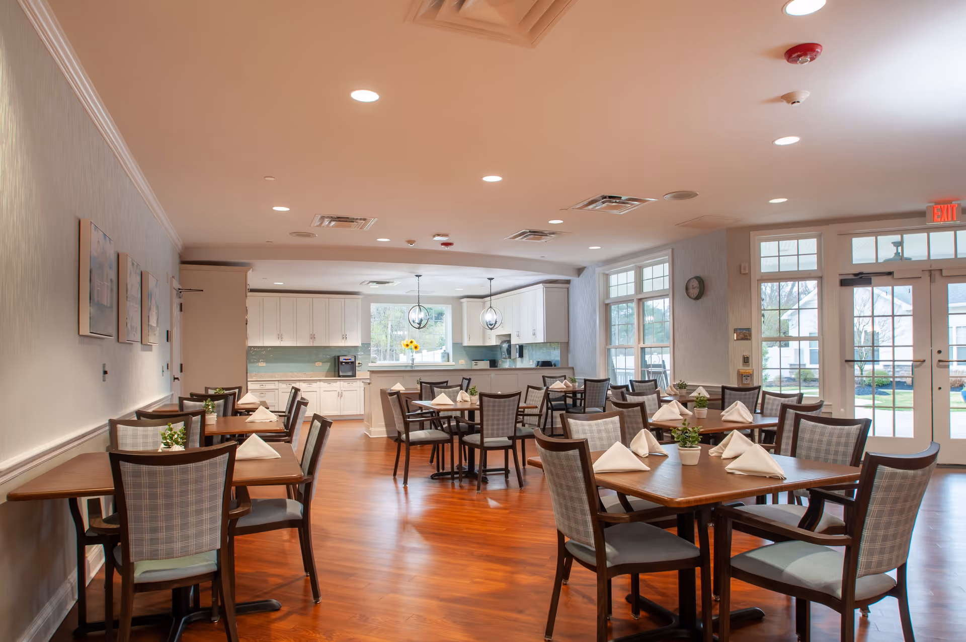 A bright and spacious dining room in Arbor Terrace Willistown featuring multiple wooden tables with chairs arranged neatly around them. Each table is set with folded white napkins and small potted plants. The room has large windows allowing natural light to fill the space, and a kitchen area with white cabinets and modern lighting fixtures is visible in the background.